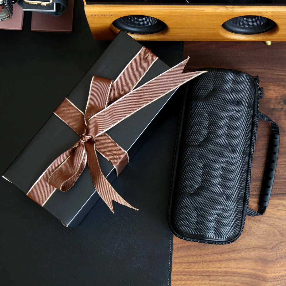 A dark leather watch travel case organizer is shown next to a wooden speaker and a gift box tied with brown ribbon.
