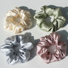 Three silk hair scrunchies in cream, sage green, and dusty rose colors arranged on a white background.