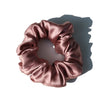 A close-up of a ruched, satin hair scrunchie in a dusty rose color.