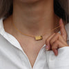A close-up of a gold plated rectangle pendant necklace worn around the neck against a white shirt.