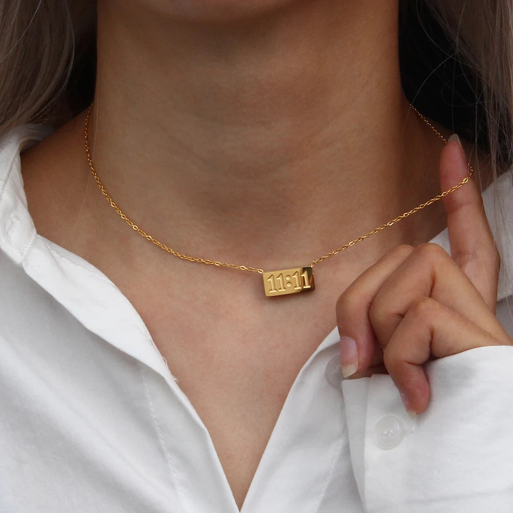 A close-up of a gold plated rectangle pendant necklace worn around the neck against a white shirt.