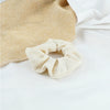 A beige knitted hair scrunchie headband is shown next to a white fabric on a white surface.
