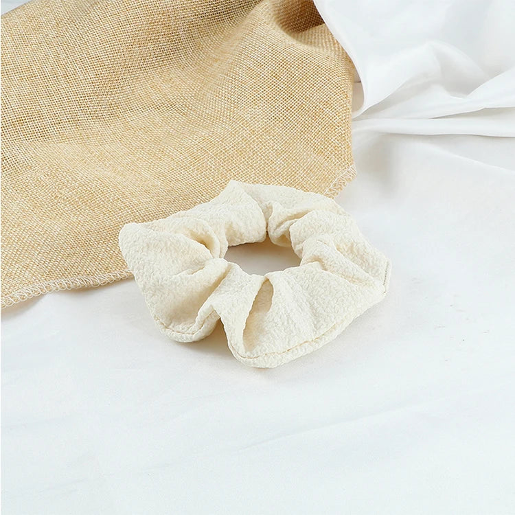 A beige knitted hair scrunchie headband is shown next to a white fabric on a white surface.