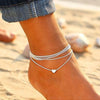 Silver and gold heart chain anklets worn on a woman's ankle against a sandy background.