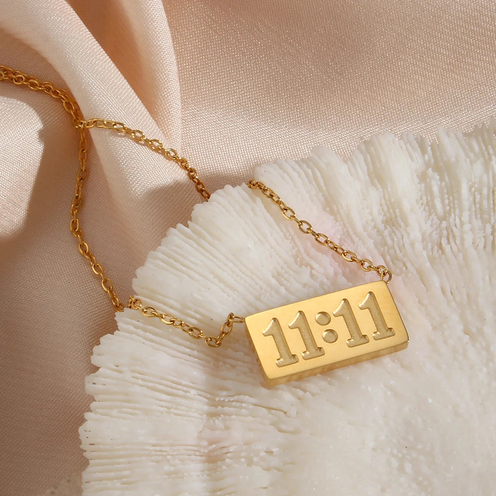 A gold-plated rectangle pendant necklace showing the time 11:11 resting on a white, textured surface.