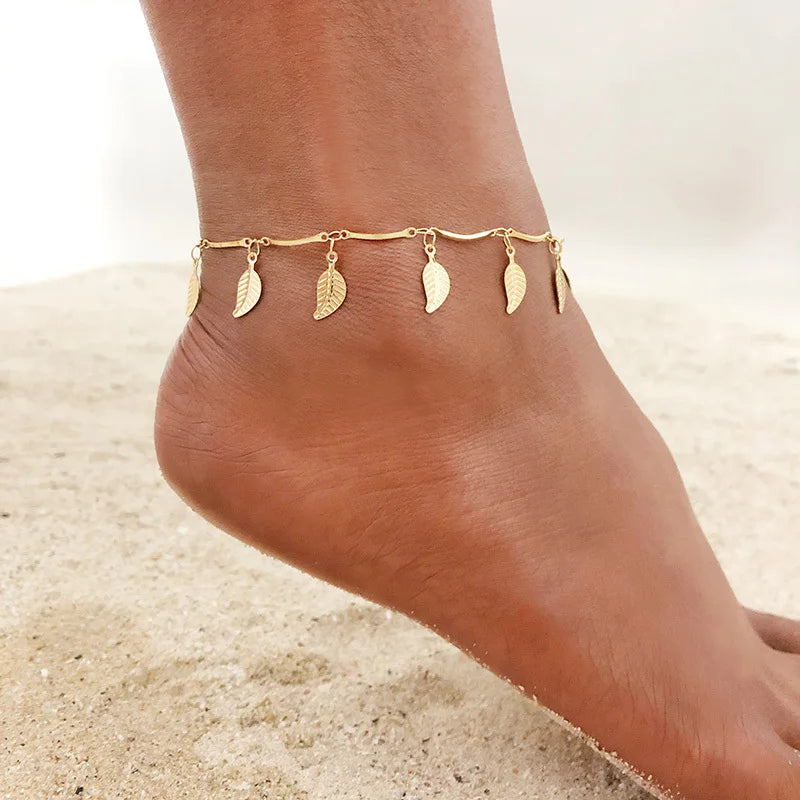 Gold stainless steel anklets with leaf charms worn on a woman's ankle against a sandy background.