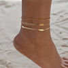 Gold and silver heart chain anklets worn around a woman's ankle on a sandy surface.