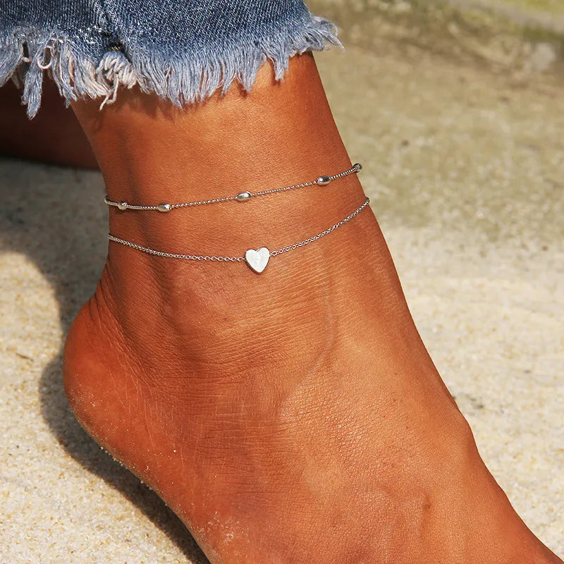 Silver and gold heart chain anklets worn on a woman's ankle against a sandy background.
