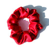A vibrant red silk hair scrunchie with gathered fabric texture casting a shadow on a white surface.