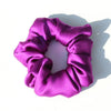 A purple silk hair scrunchie is shown lying on a white surface.