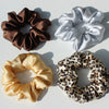 Three silk hair scrunchies in brown, silver, and gold satin fabric are displayed on a white background.
