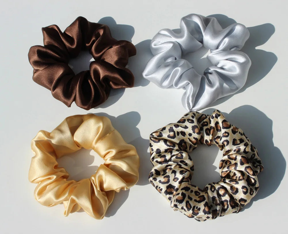 Three silk hair scrunchies in brown, silver, and gold satin fabric are displayed on a white background.