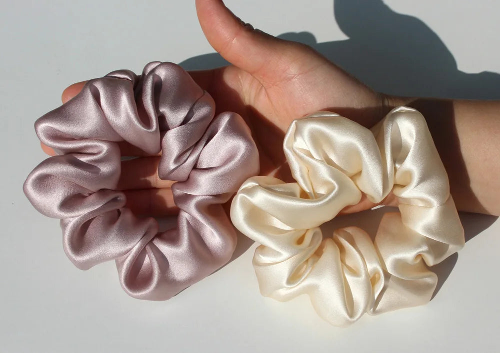 A hand holds a pair of light pink and cream silk hair scrunchies against a white background.