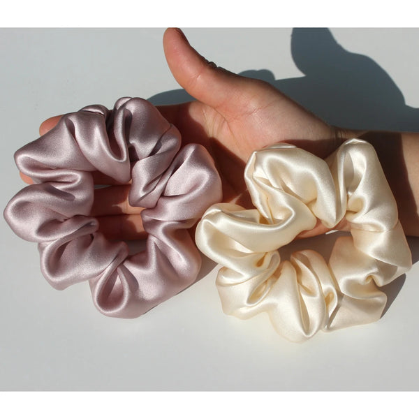 A hand holds two silk hair scrunchies, one in dusty rose and one in cream.