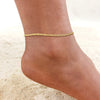 A close-up of a gold stainless steel anklet worn around a woman's ankle on a sandy surface.
