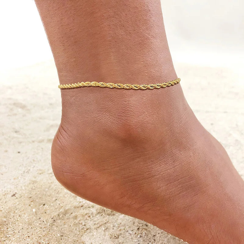 A close-up of a gold stainless steel anklet worn around a woman's ankle on a sandy surface.