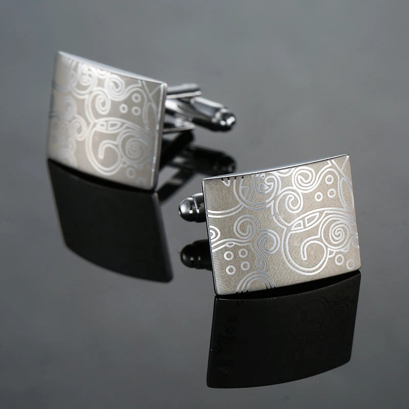 Silver square cufflinks with an engraved swirling pattern displayed on a black surface.