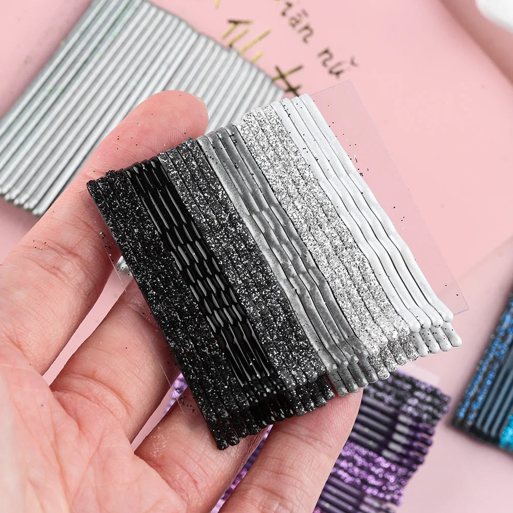 A hand holds a set of 24 glitter wavy hair clips and bobby pins in various shades of black, silver, and purple.