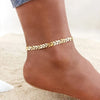 Gold stainless steel anklet worn on a woman's ankle against a sandy background.