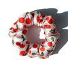 A circular scrunchie made of white silk fabric with red and green cherry print.