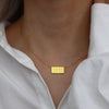 A close-up of a gold plated rectangle pendant necklace with the time 11:11 on a delicate chain, worn against a white shirt.