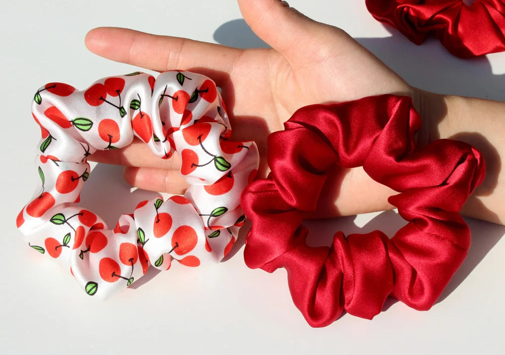 A hand holds two sets of luxury pure silk hair scrunchies, one white with red fruit designs and one solid red.