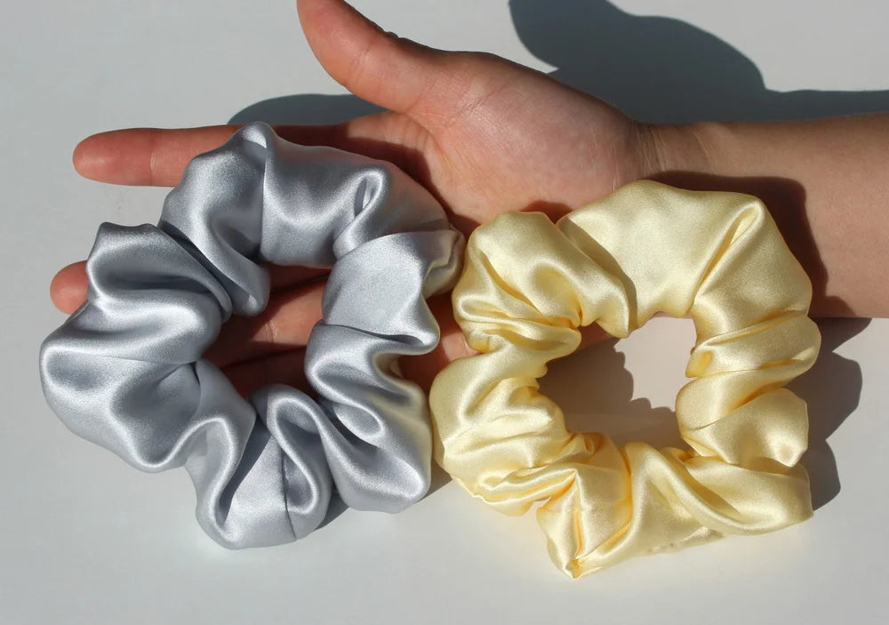 A pair of luxury pure silk hair scrunchies in silver and pale yellow are held by a hand.