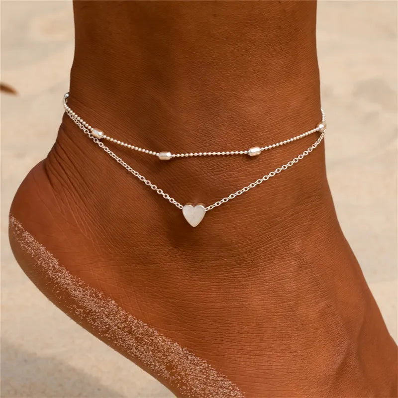 Silver heart chain anklets worn on a woman's ankle against a sandy background.