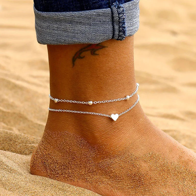 Silver and gold heart chain anklets worn on a foot against a sandy background.