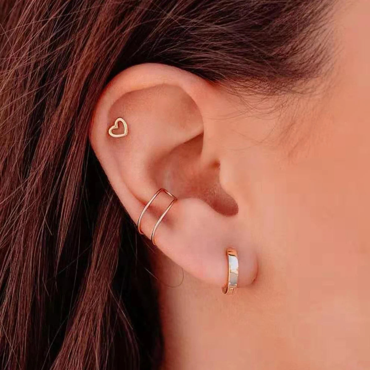 Close-up of a woman's ear showing minimalist gold hoop earrings and a small heart stud.