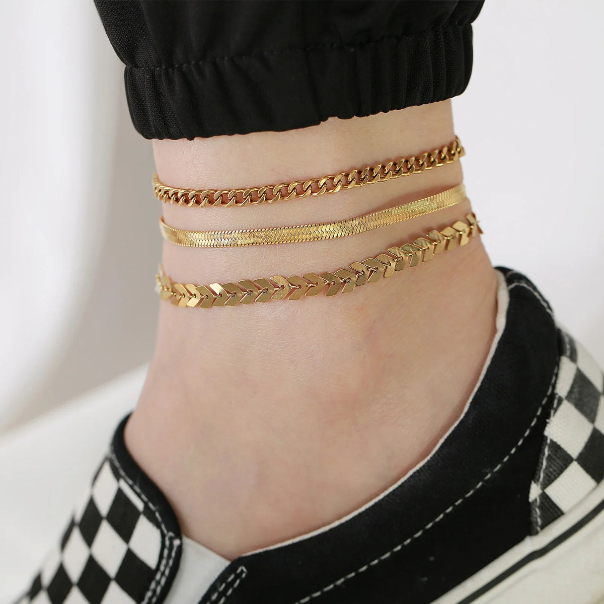 Gold stainless steel anklets worn around the ankle of a person wearing black clothing and sneakers.
