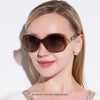 Eine Frau präsentiert klassische braune Sonnenbrille mit goldenen Akzenten vor weißem Hintergrund.