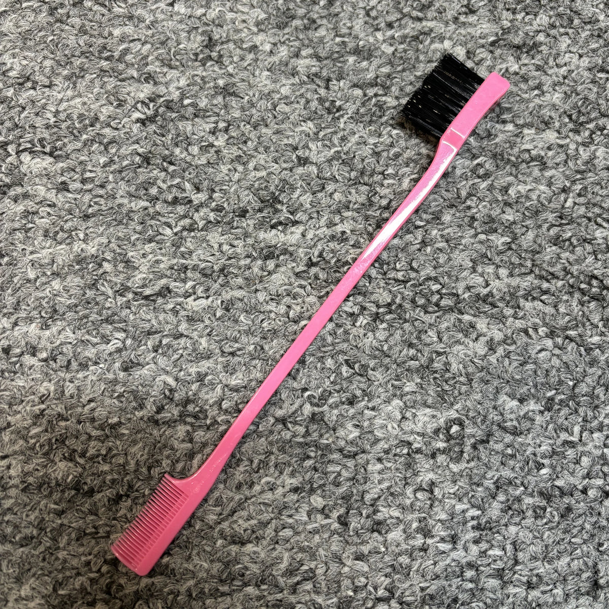 Pinkes, zweischneidiges Augenbrauenkamm mit schwarzen Borsten auf grauer, strukturierter Oberfläche.