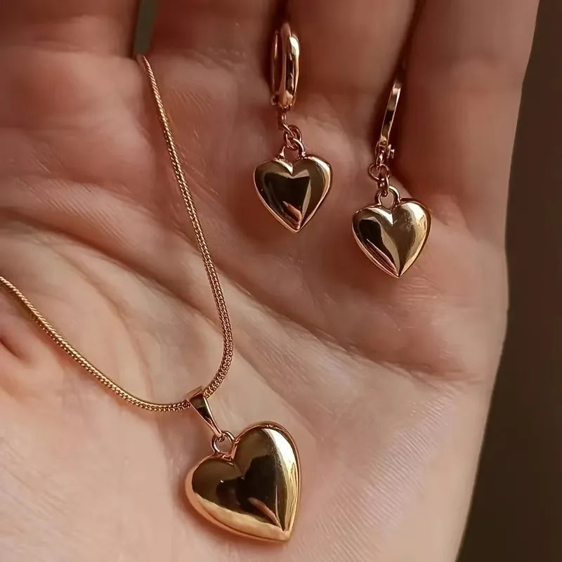 A close-up of a delicate gold heart necklace and matching earring set displayed on a person's hand.