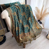 A dark teal and gold bohemian paisley tassel shawl draped over a light-colored cushion.