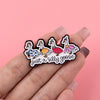A hand holds a set of black enamel pins depicting a silly goose character.