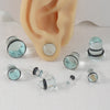 A glass ear stretching kit showing various sized ear plugs and tunnels next to a light peach-colored ear model.
