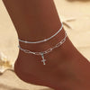 A close-up of a sparkling zircon heart cross anklet worn on a person's ankle against a sandy background.