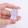 A hand holds a purple enamel pin shaped like a ribbon with the text Got Out Of Bed Today Yay and a yellow star.