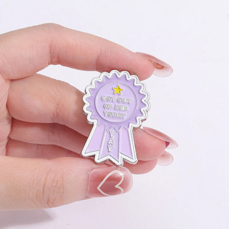 A hand holds a purple enamel pin shaped like a ribbon with the text Got Out Of Bed Today Yay and a yellow star.