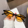A hand holds a pair of retro oval frame sunglasses with amber lenses against a light background.