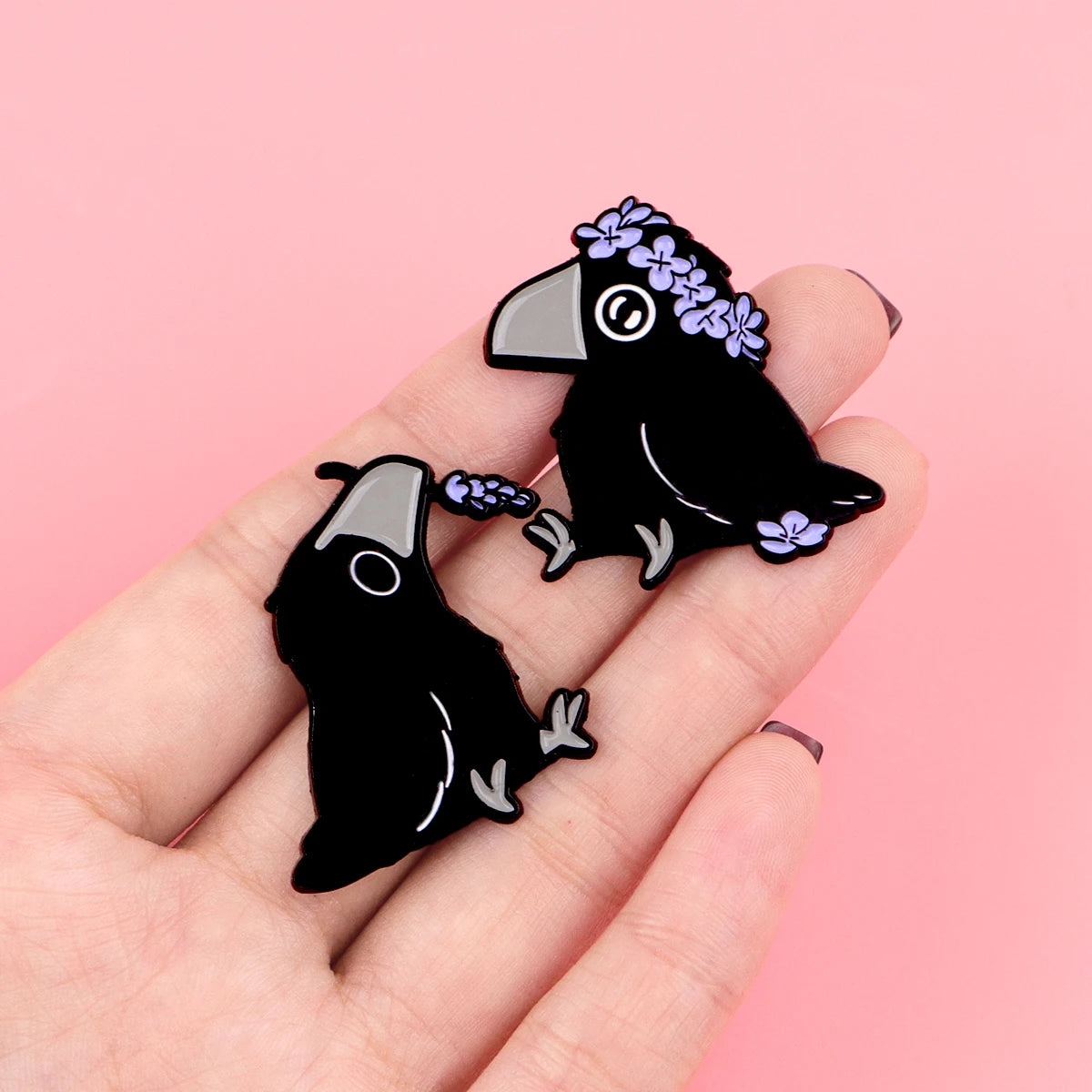 Eine Hand hält eine schwarze Emaille-Anstecknadel mit einer Krähe und lila Blumen.