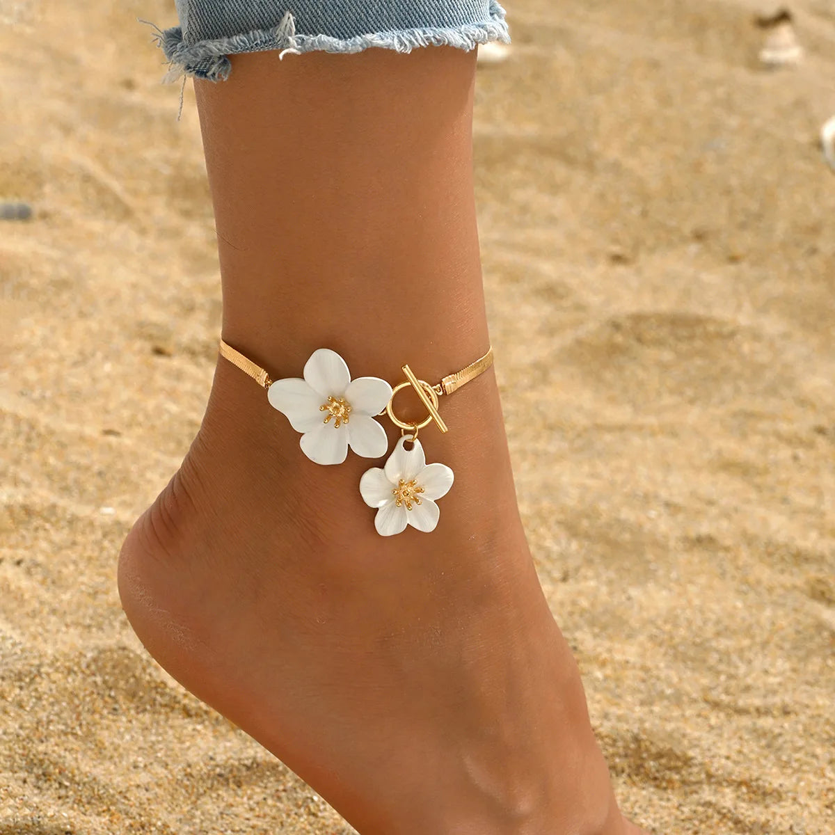 A gold chain anklet featuring white flower pendants worn on a person's ankle in a sandy outdoor setting.