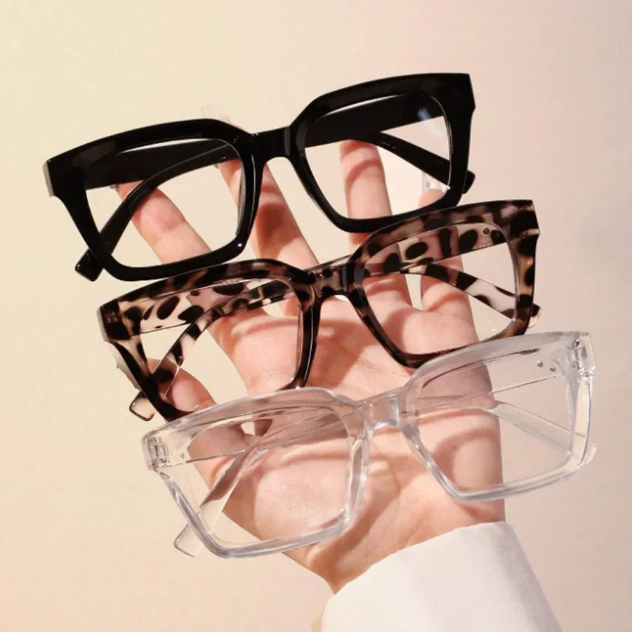 A hand holds a pair of black, tortoiseshell-patterned square reading glasses against a light background.