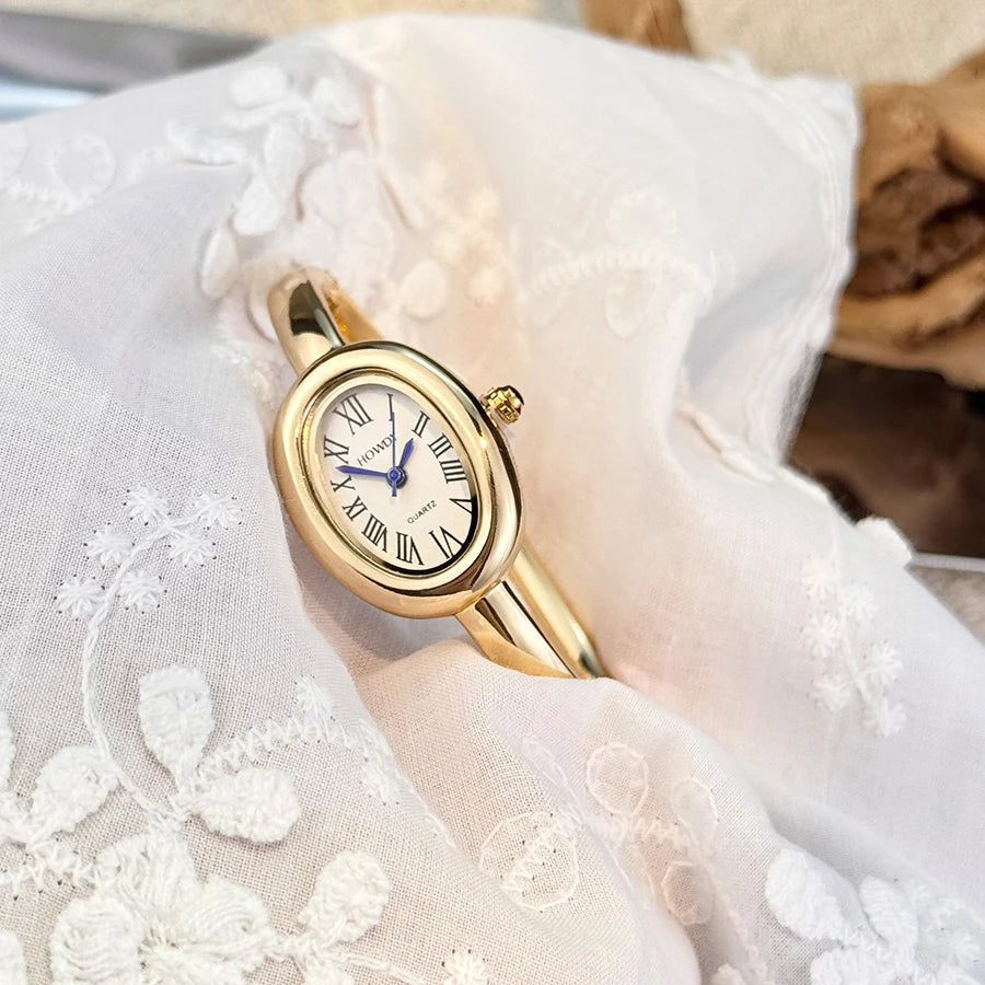 A gold-toned quartz watch with a white face and Roman numerals is displayed on sheer white fabric.