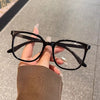 A hand is holding a pair of black, classic square anti-blue light eyeglasses against a light gray background.