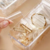A hand is opening a clear self-sealing jewelry storage pouch revealing several gold-toned rings and earrings inside.