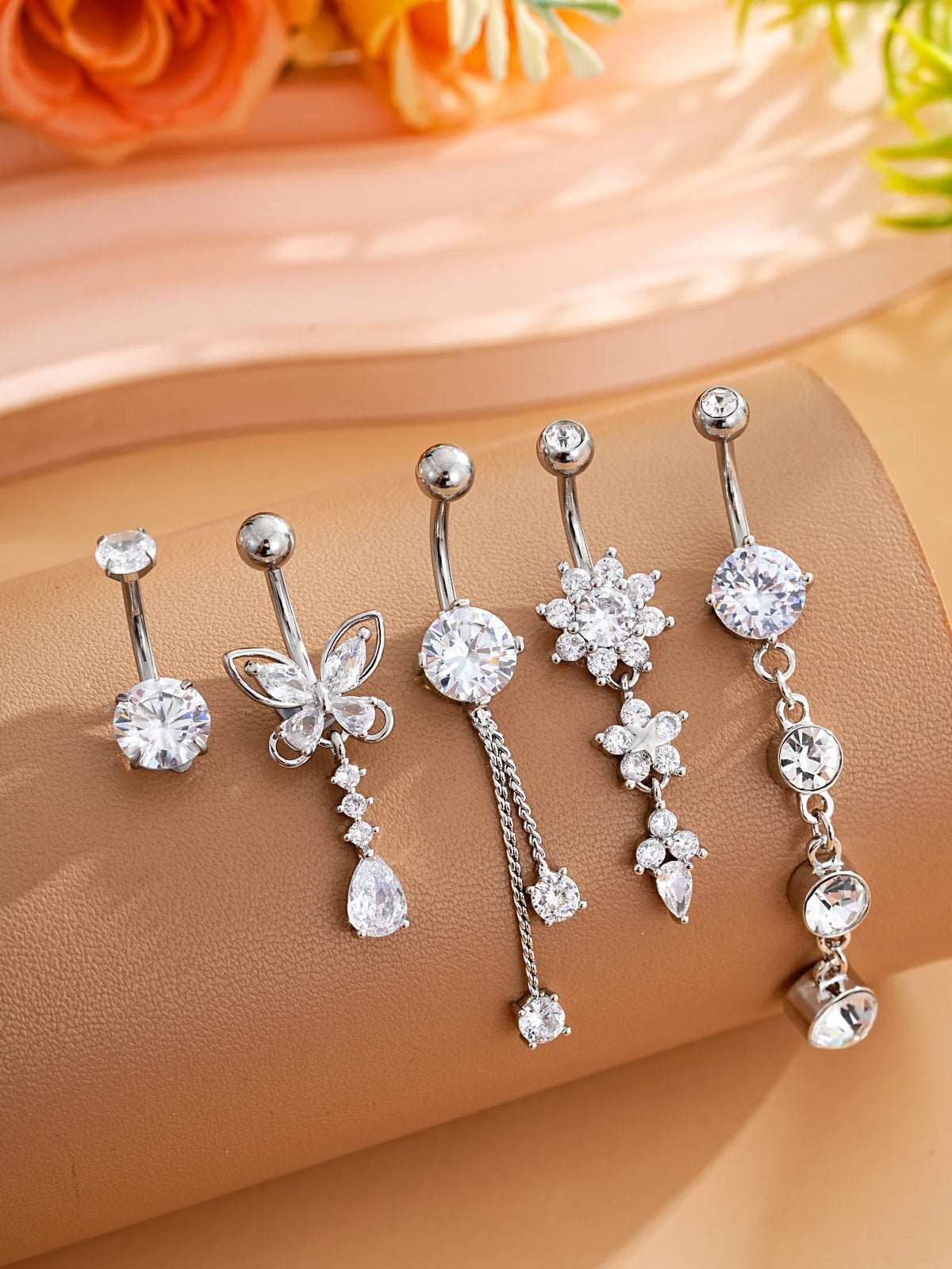 A set of five gold stainless steel dangled belly button rings with various diamond and floral designs displayed on a beige su