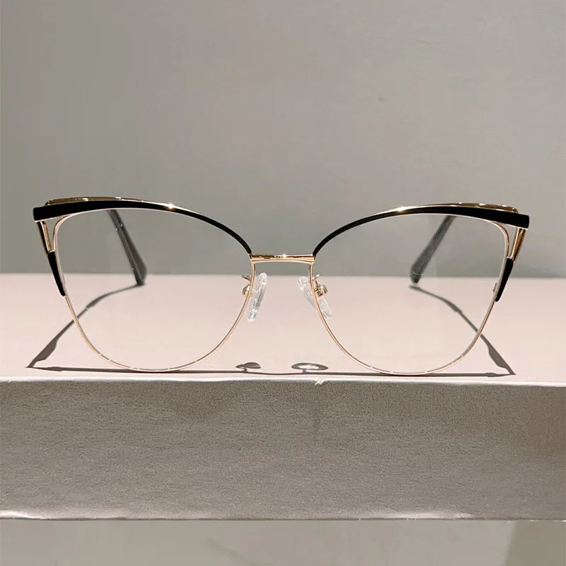 Un par de gafas de computadora con montura negra y efecto ojo de gato de metal dorado sobre una caja blanca.