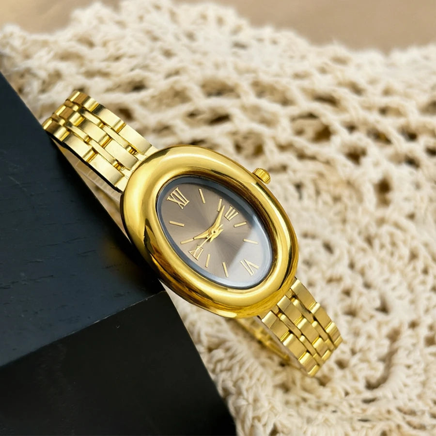 Reloj vintage dorado con esfera clara y correa de metal sobre una superficie oscura junto a tela tejida beige.